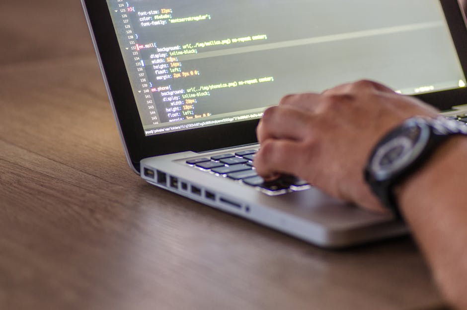 A person working on a laptop placed on a wooden surface, with visible code on the screen. The individual's right hand, wearing a dark wristwatch, is resting on the keyboard, suggesting active input or
